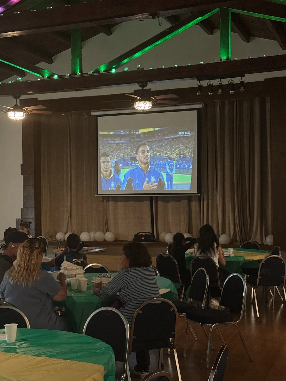 Guests eating dinner at green-covered tables with the 2026 World Cup match on the projector screen